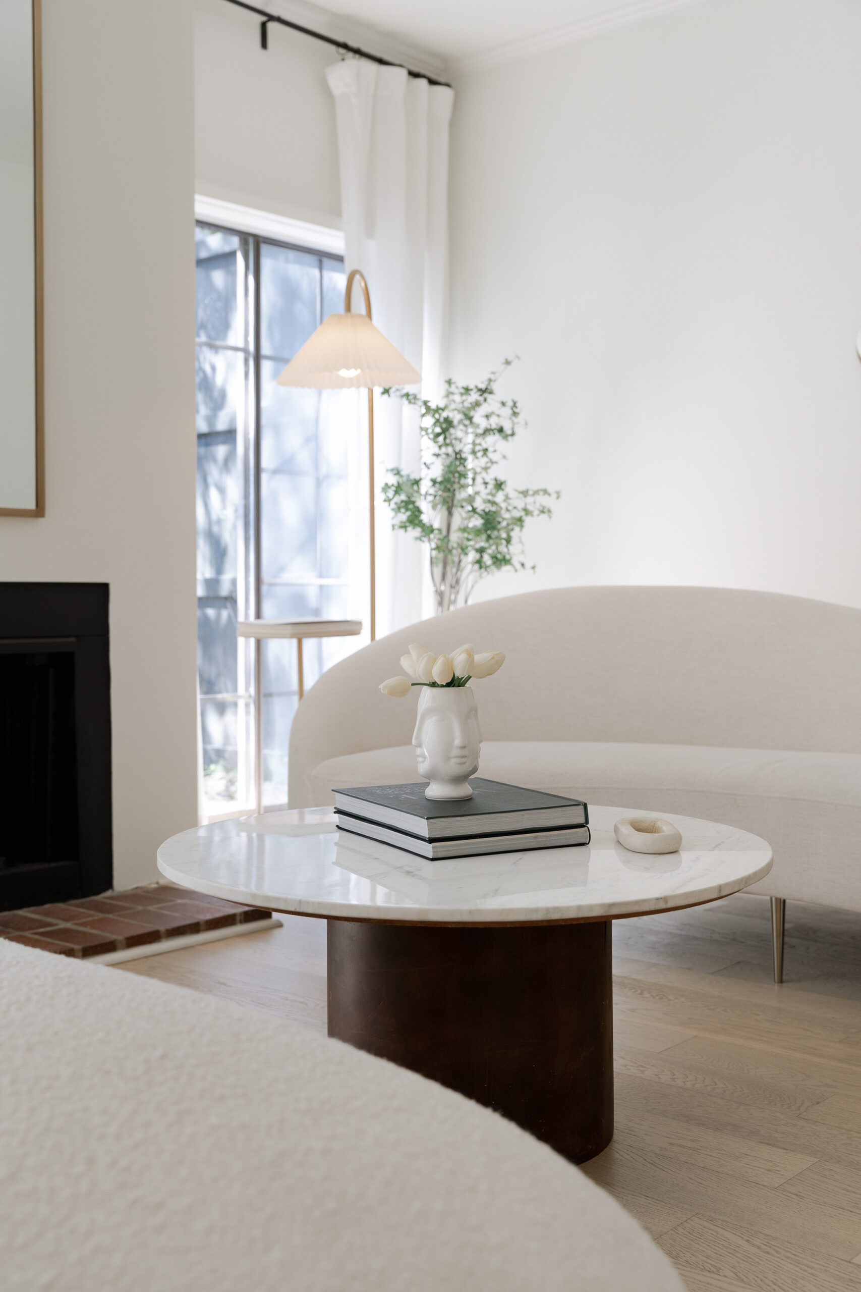 Peaceful living room with soft natural light, curved sofa, marble coffee table with books and white tulips, and greenery by the window