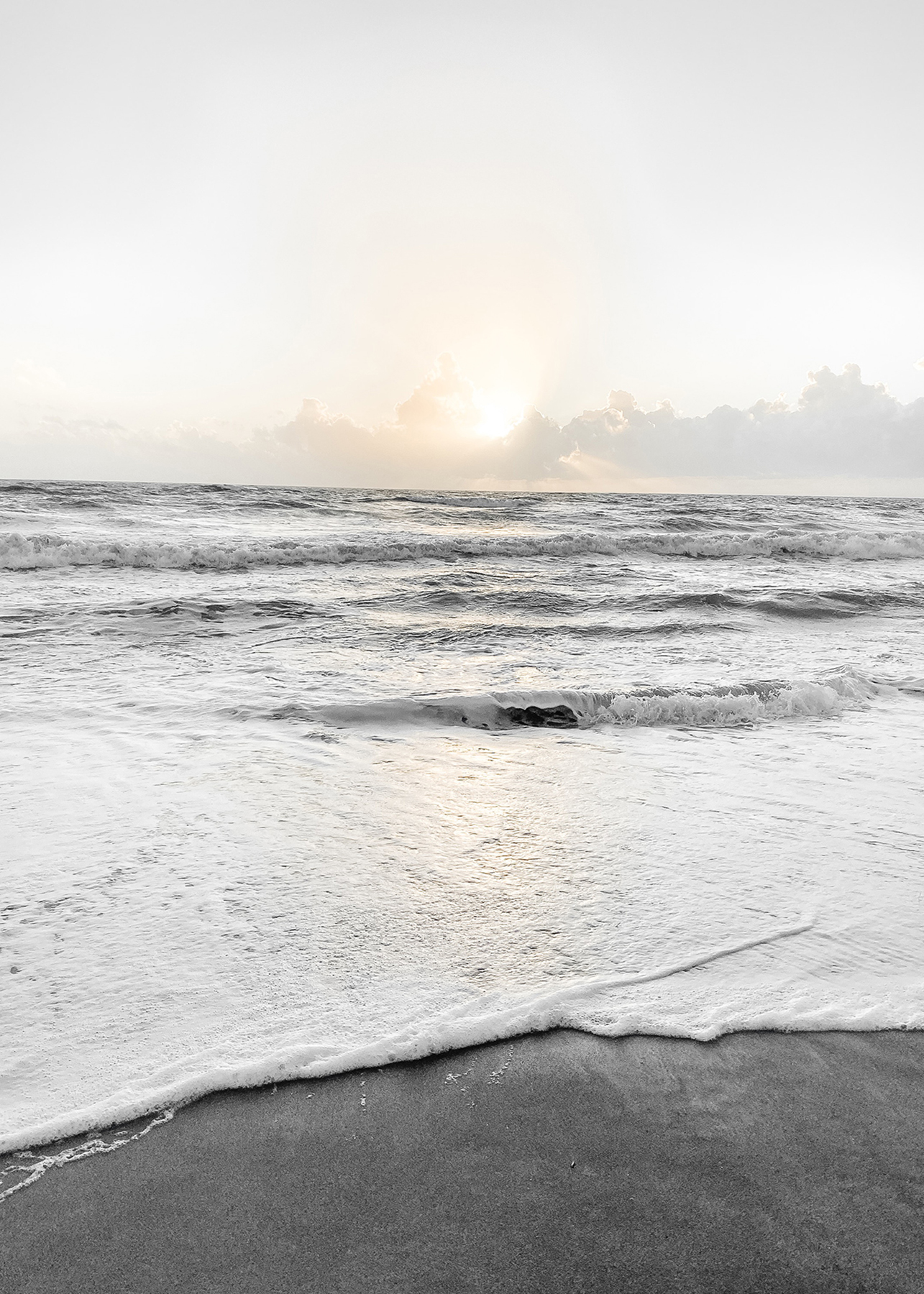 Gentle ocean waves washing onto a sandy beach at sunrise, with soft golden light breaking through clouds on the horizon