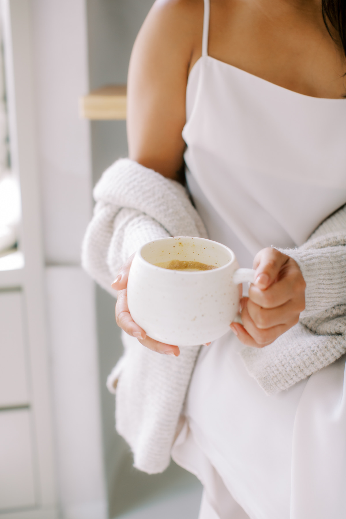Woman in soft cardigan gently cradling a warm mug in both hands, soft morning light in the background
