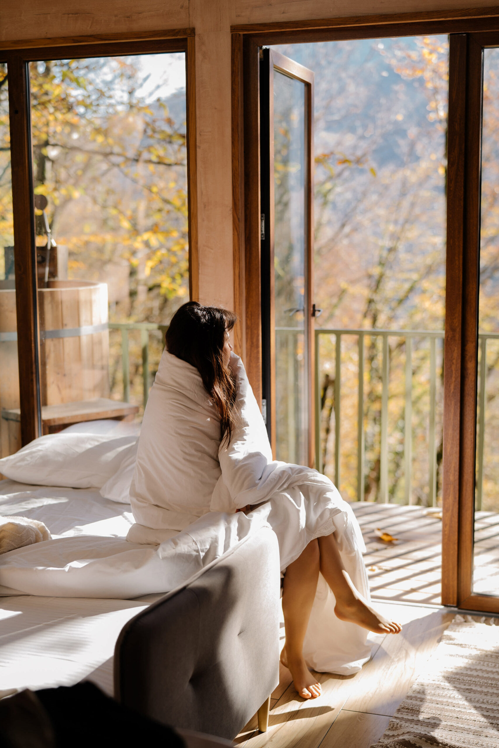 A woman with long dark hair sits on the edge of a bed, wrapped cozily in a white duvet, looking out through floor-to-ceiling glass doors at a balcony surrounded by trees in golden autumn foliage. Warm morning sunlight streams through the windows, casting gentle shadows on the light wooden floor. The serene cabin setting features natural wood framing and creates an atmosphere of peaceful contemplation and rest.